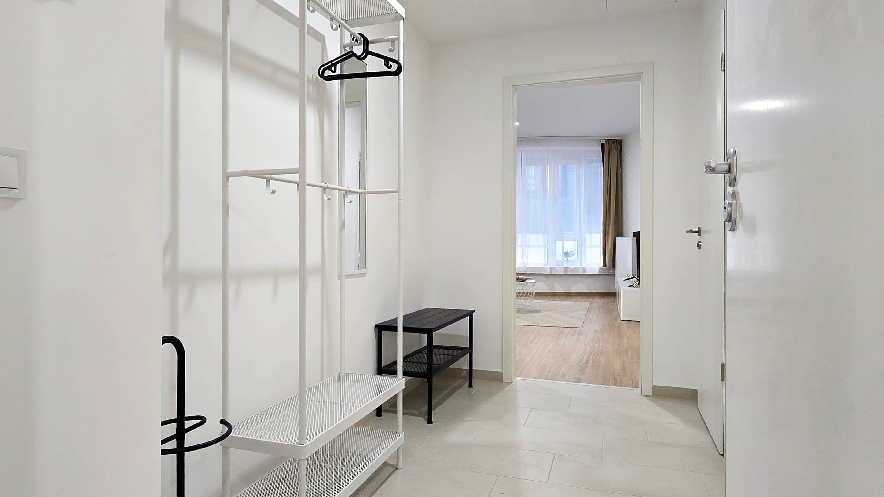 Un couloir avec une porte blanche, un banc noir, une étagère blanche et un cintre noir.