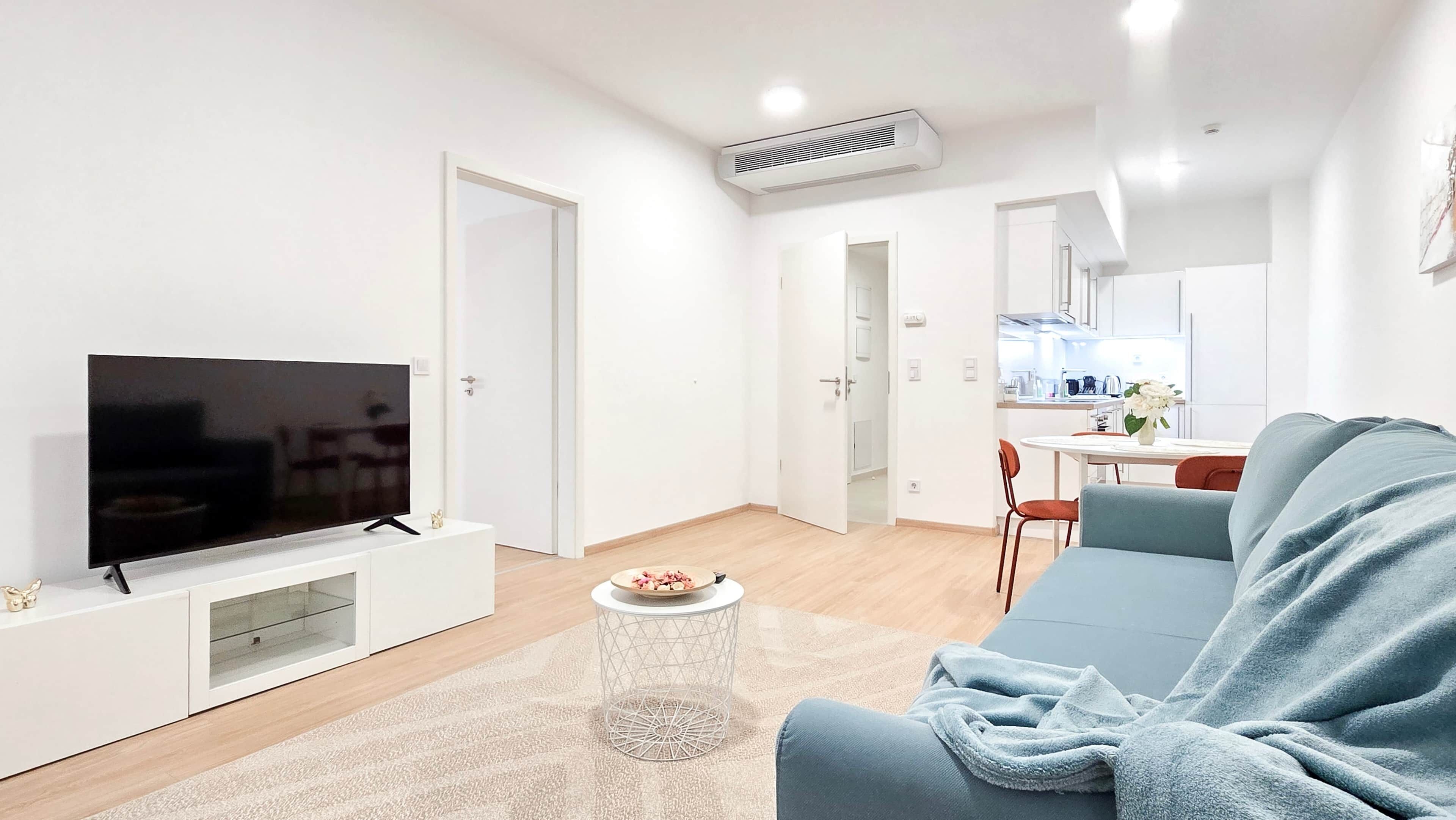 A modern living room with a flat-screen TV, a sofa, a coffee table, and a kitchen area in the background.