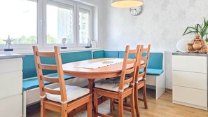 A dining area with a round table and wooden chairs, a clock on the wall, and a window with blinds. - LB - Apartman Mabelu (Siófok)