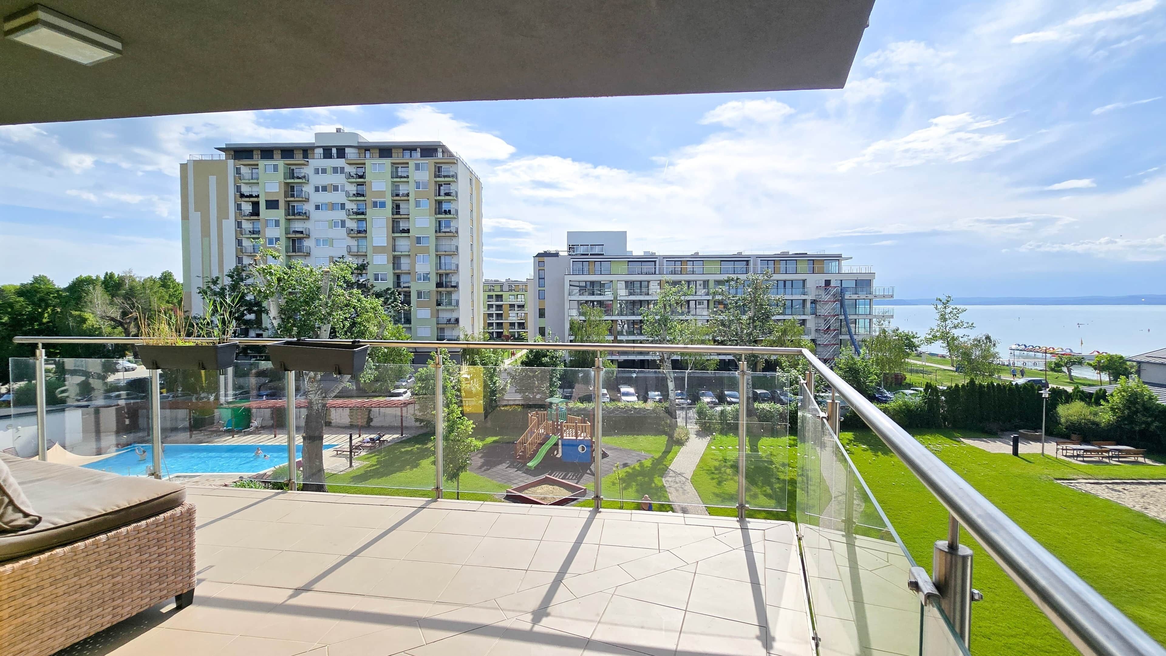 A balcony with a view of a swimming pool, a grassy area, and buildings in the background.