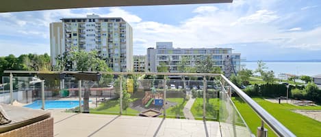 A balcony with a view of a swimming pool, a grassy area, and buildings in the background.