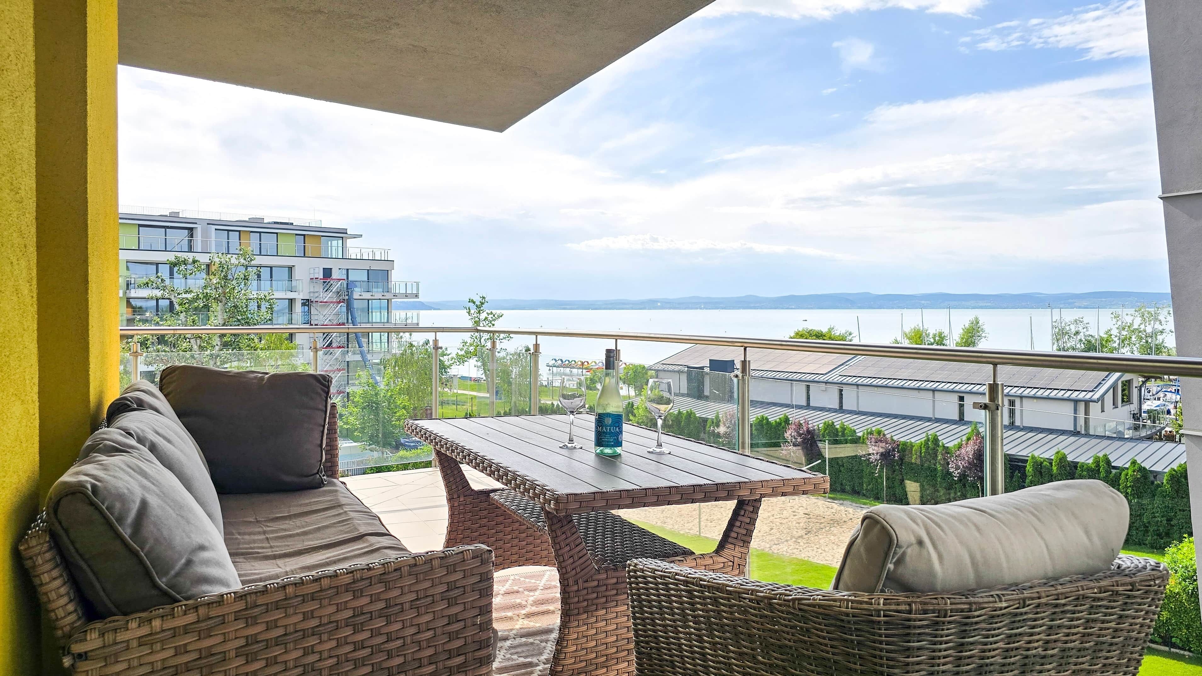 A balcony with wicker furniture, a table, and two glasses, overlooking a body of water and buildings.