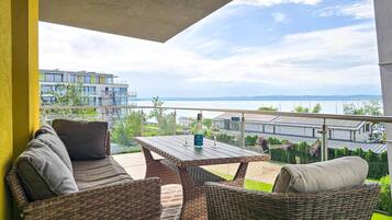 A balcony with wicker furniture, a table, and two glasses, overlooking a body of water and buildings.