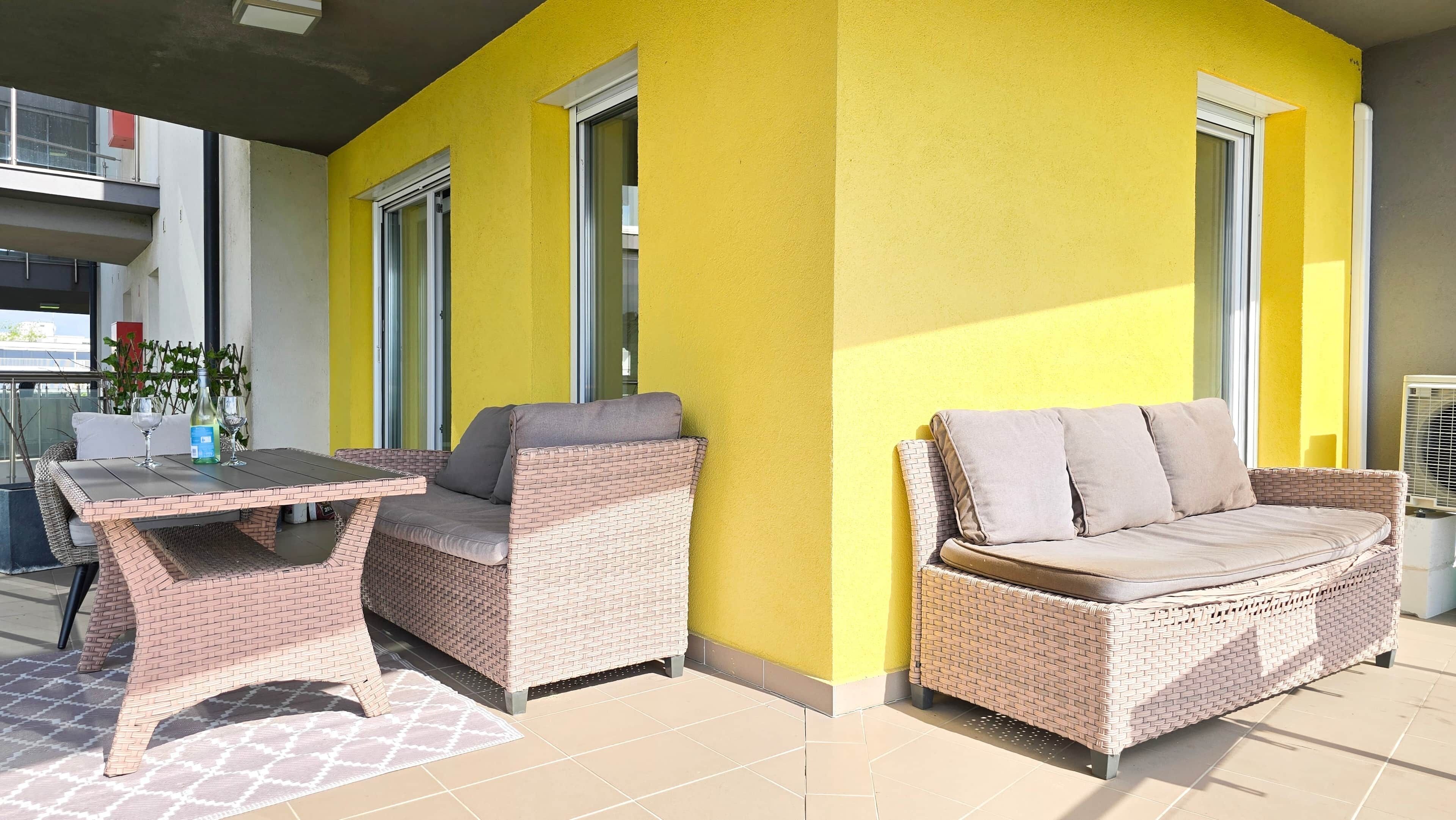 An outdoor seating area with wicker furniture and a table, adjacent to a yellow wall with windows.