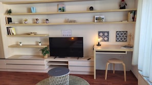 A living room with a flat-screen TV, shelves with books and decorative items, a desk with a chair, and a small round table. - IMCT - Passage Apartman (Budapest)