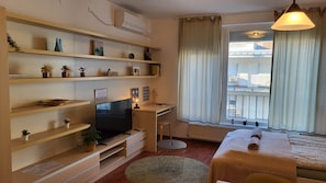 A modern living room with a TV, shelves with plants, a dining area with a table, and a window with curtains. - IMCT - Passage Apartman (Budapest)