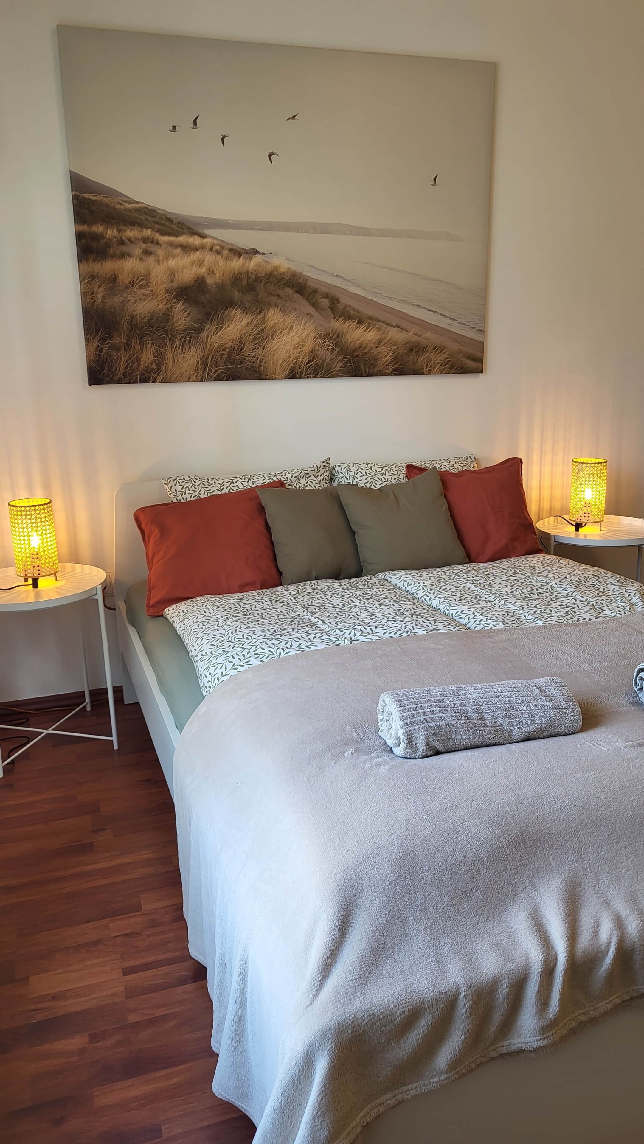 Un dormitorio con cama, almohadas, una colcha, una toalla, una mesita de noche con lámpara y un cuadro de aves colgado en la pared.