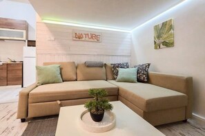 Ein modernes Wohnzimmer mit einer Eckcouch, einem Couchtisch mit Pflanze, einer Küchenzeile mit Mikrowelle und einem an der Wand befestigten Schild mit der Aufschrift 'NATURE'.