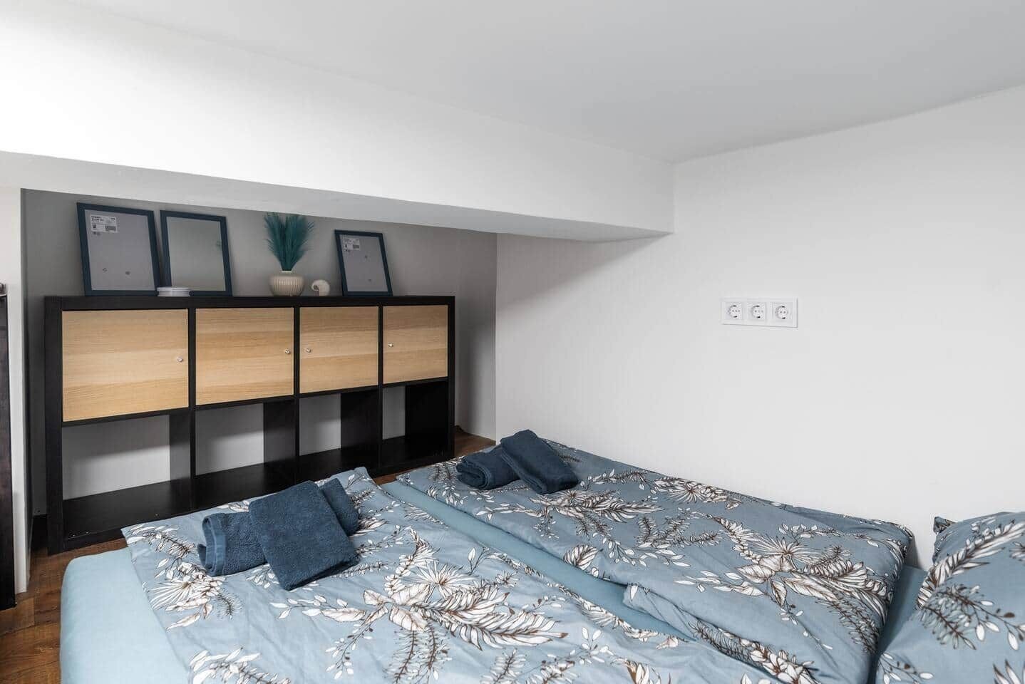 A bedroom with a bed, a nightstand, a dresser, and a wall-mounted shelf with framed pictures.
