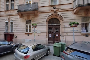 A building with a central entrance, two balconies, and a parking area with cars and green bins. - IMCT - Rose Apartment mini (Budapest)