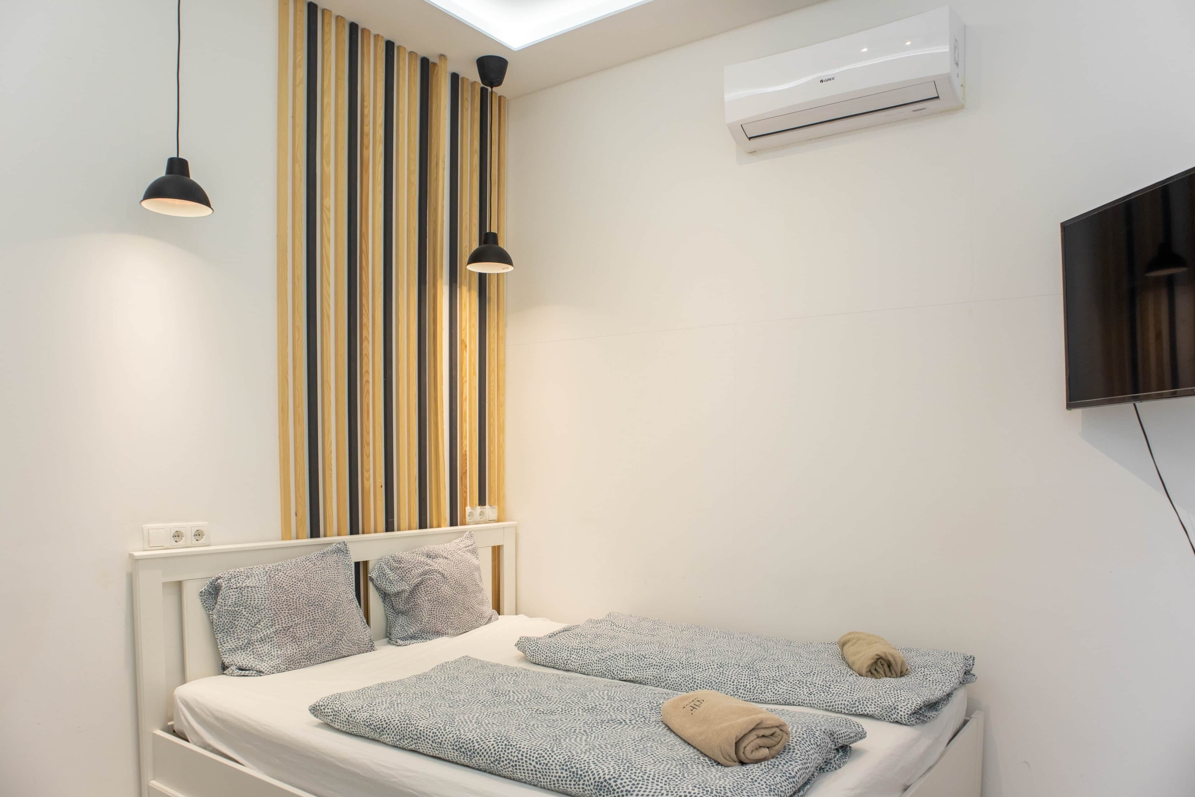 A bedroom with a bed, two pillows, a bedside table, a wall-mounted air conditioner, and a TV.