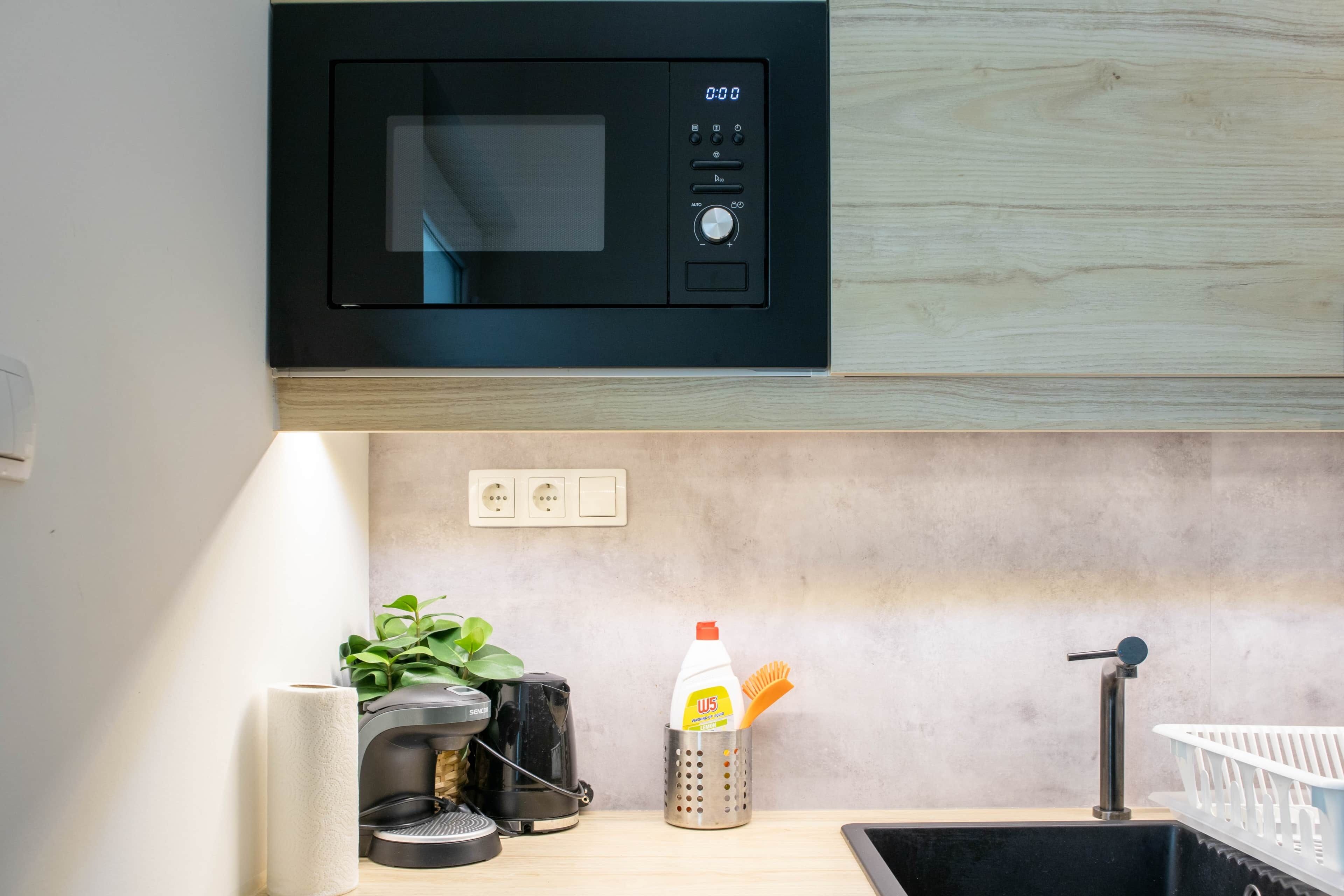 A kitchen with a black microwave, a coffee machine, a bottle of liquid soap, and a white basket.
