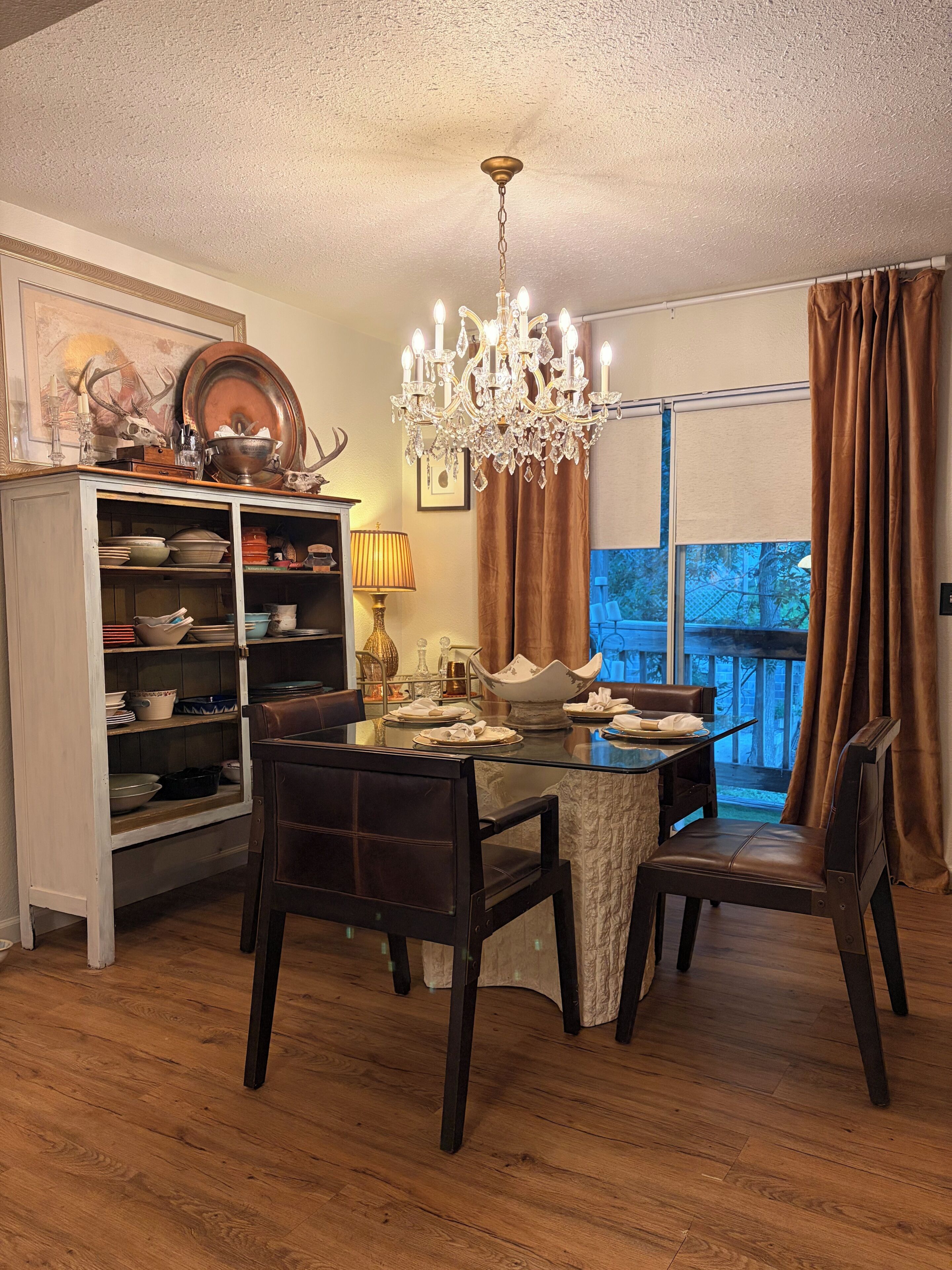 A dining area with a glass table, dark chairs, a chandelier, a cabinet with dishes, and a window with curtains.