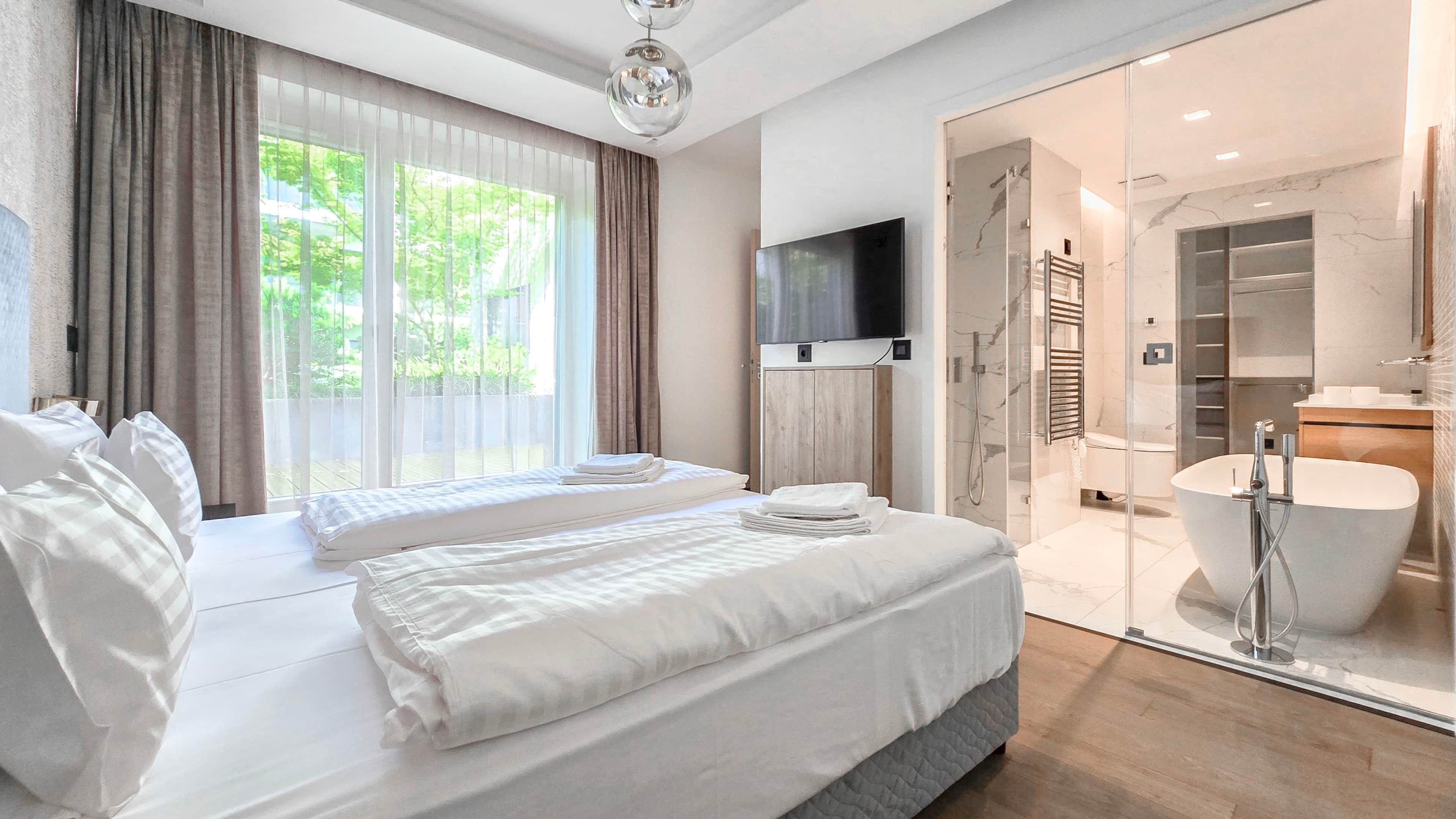 A modern hotel room with a large bed, a television, a glass-enclosed bathroom with a bathtub, and a view of greenery outside.