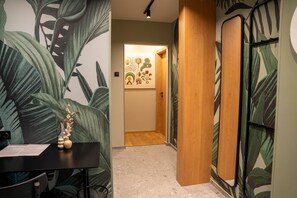 A hallway with a wooden door, a wall with a tropical leaf mural, and a small table with a vase.