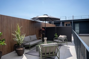 A modern outdoor seating area with a sofa, chairs, and a table, surrounded by potted plants and a large umbrella. - IMCT - Urban Jungle Apartments 06 (Budapest)