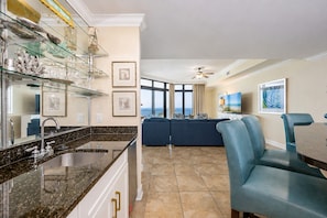 A modern kitchen with a granite countertop, a built-in sink, and a dining area with blue upholstered chairs and a table. - The Oasis 1604 Formerly Phoenix West Ii 1604 3 Bedroom Condo (Orange Beach)