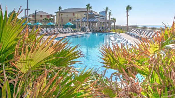 A pool area with lounge chairs and umbrellas, surrounded by palm trees and a building with a glass roof. - The Oasis 1604 Formerly Phoenix West Ii 1604 3 Bedroom Condo (Orange Beach)