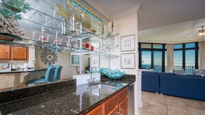 A modern kitchen with a granite countertop, a glass display shelf, and a dining area with blue furniture. - The Oasis 1604 Formerly Phoenix West Ii 1604 3 Bedroom Condo (Orange Beach)
