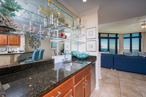 A modern kitchen with a granite countertop, a glass display shelf, and a dining area with blue furniture. - The Oasis 1604 Formerly Phoenix West Ii 1604 3 Bedroom Condo (Orange Beach)