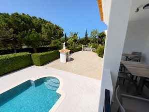 Zona de piscina con solárium de baldosas, una pequeña construcción blanca y vista a una zona vallada con árboles.