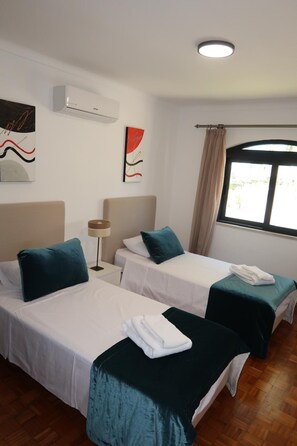 A hotel room with two single beds, a bedside table with a lamp, a window with curtains, and a wall-mounted air conditioner. - Villa Irene 7 Bed Villa close to Marina (Vilamoura)