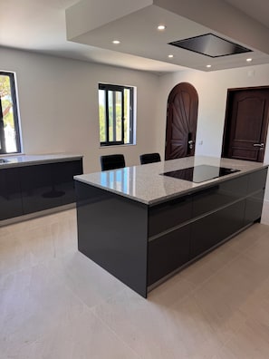 A modern kitchen with a large island, dark cabinetry, and a window with blinds. - Villa Irene 7 Bed Villa close to Marina (Vilamoura)