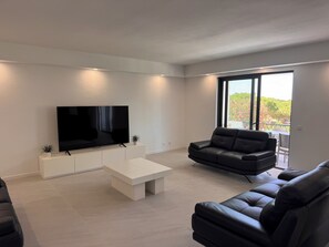 A modern living room with a flat-screen TV, a sofa, a coffee table, and large windows offering a view of greenery. - Villa Irene 7 Bed Villa close to Marina (Vilamoura)