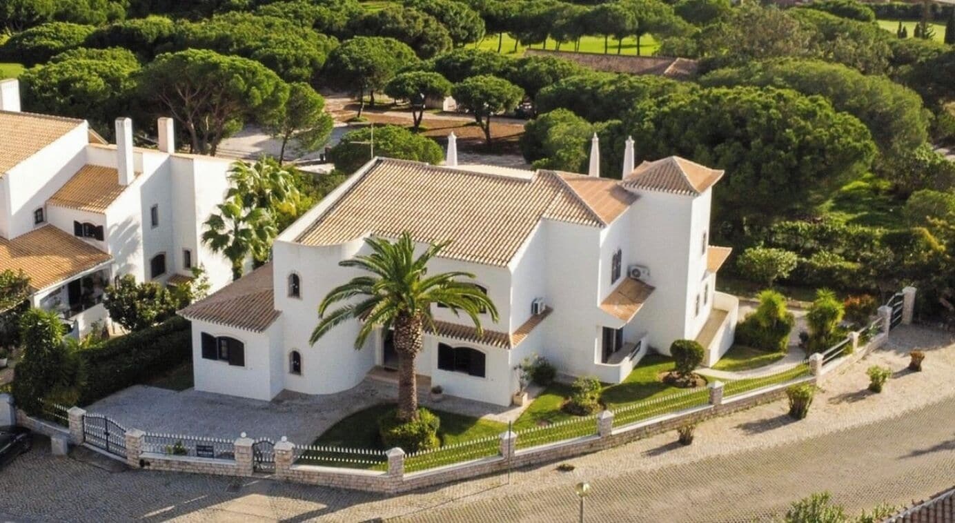 A white villa with a tiled roof, surrounded by lush greenery and a paved driveway.