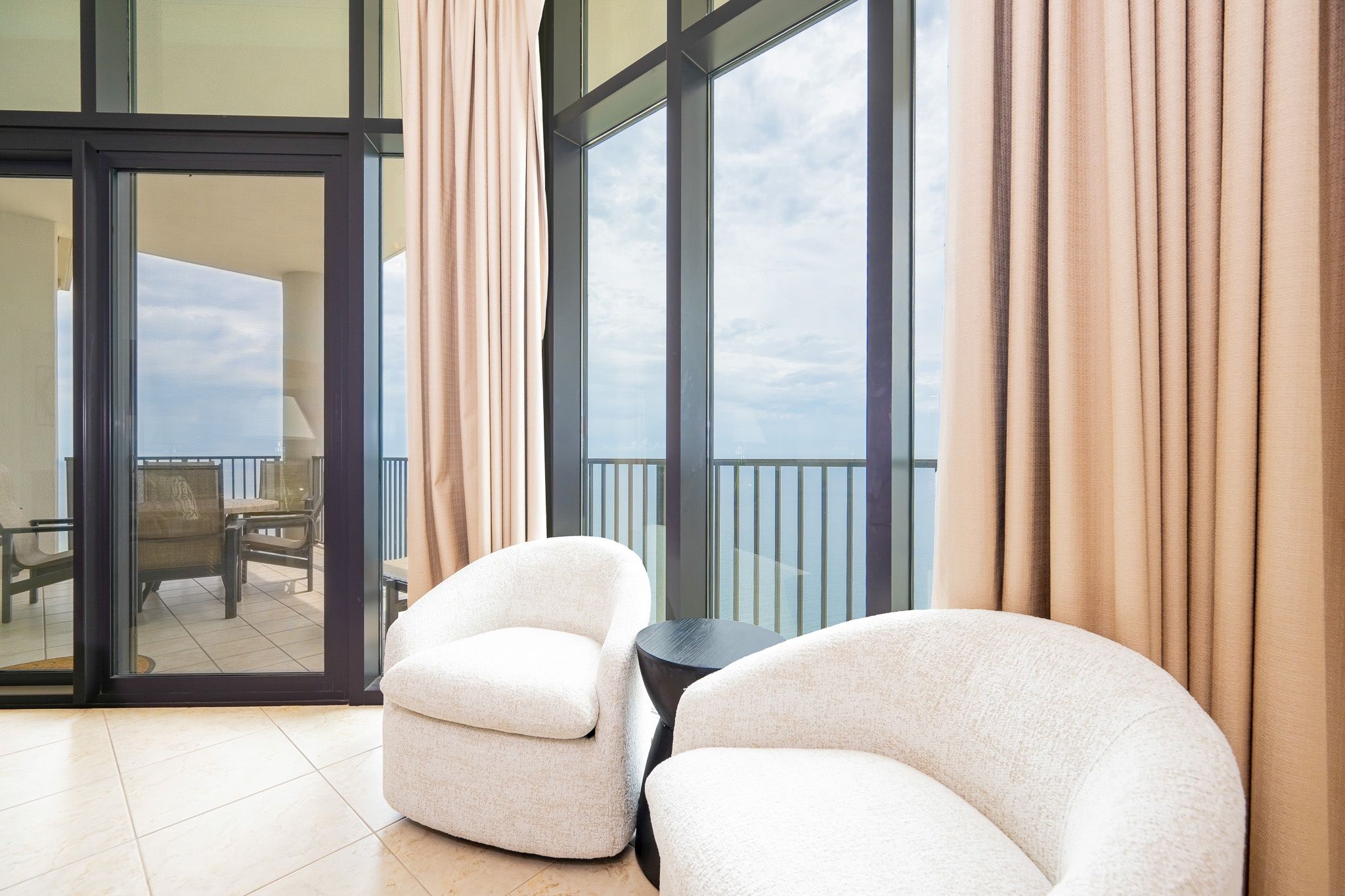 A modern room with two plush armchairs, a small round table, and a large sliding glass door leading to a balcony with a view of the sky.