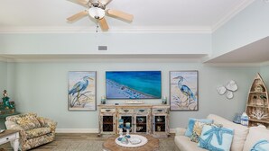 A living room with a ceiling fan, a large flat-screen TV, and three framed paintings of a beach scene. - The Oasis 3003 Formerly Phoenix West Ii 3003 3 Bedroom Condo (Orange Beach)