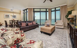 A spacious living room with a large window, a brown leather sofa, a beige armchair, and a patterned ottoman. - The Oasis 503 Formerly Phoenix West Ii 503 3 Bedroom Condo (Orange Beach)