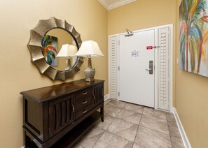 A hotel lobby with a dark wooden console, a round mirror, two lamps, a white door with a peephole, and a painting on the wall. - The Oasis 503 Formerly Phoenix West Ii 503 3 Bedroom Condo (Orange Beach)