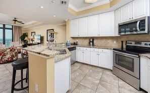 A modern kitchen with white cabinetry, a granite countertop, stainless steel appliances, and a bar area with stools. - The Oasis 503 Formerly Phoenix West Ii 503 3 Bedroom Condo (Orange Beach)