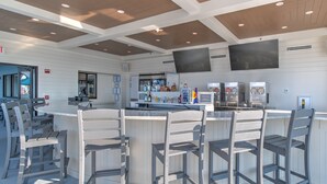 A bar area with a counter and four bar stools, a television mounted on the wall, and a refrigerator with a freezer. - The Oasis 2608 Formerly Phoenix West Ii 2608 3 Bedroom Condo (Orange Beach)