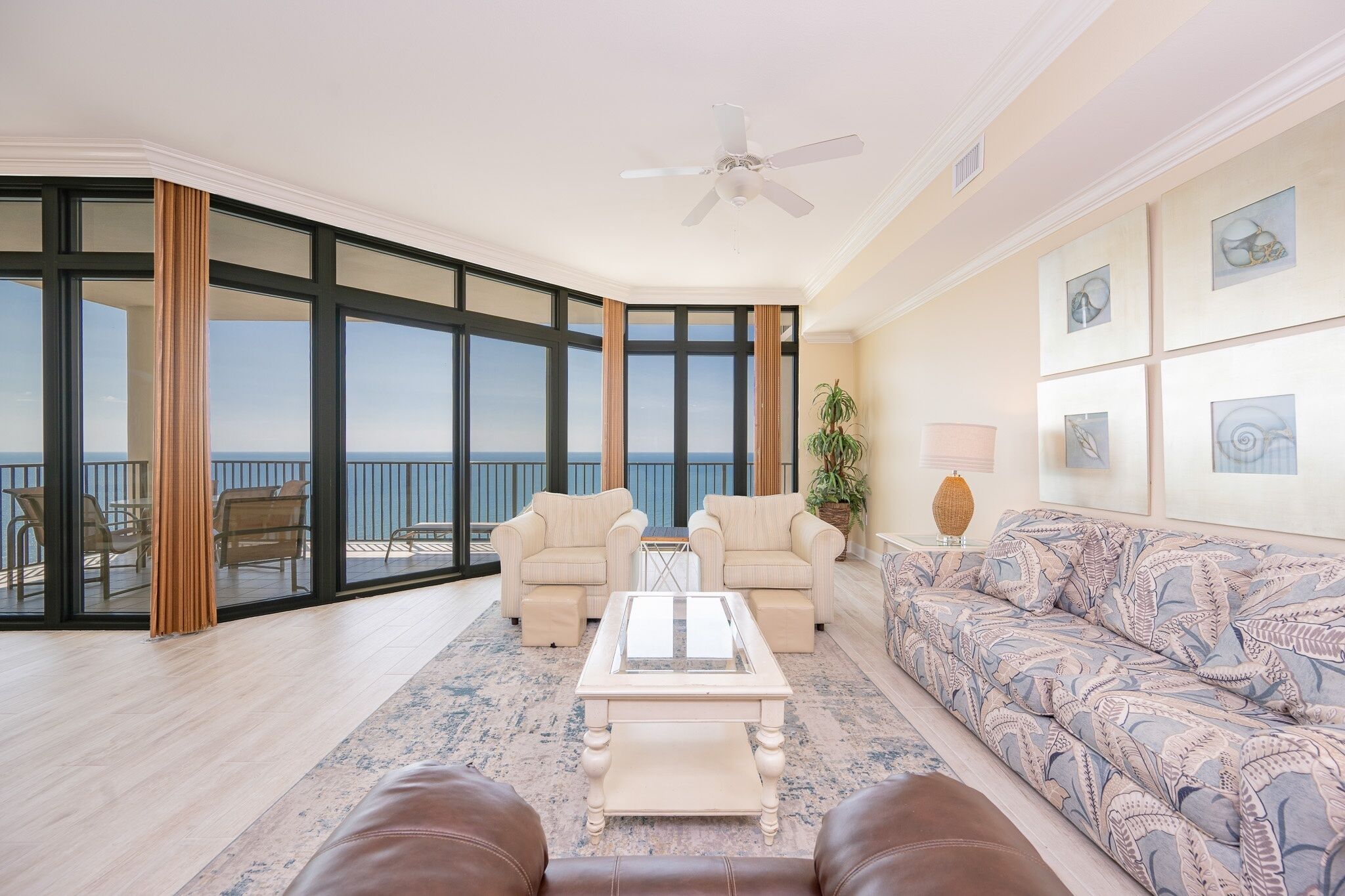 A spacious living room with a large window overlooking the sea, a ceiling fan, and a patterned sofa.