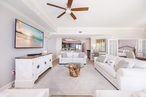 A modern living room with a ceiling fan, a large TV, and a dining area with a kitchen in the background. - The Oasis 1205 Formerly Phoenix West Ii 1205 3 Bedroom Condo (Orange Beach)