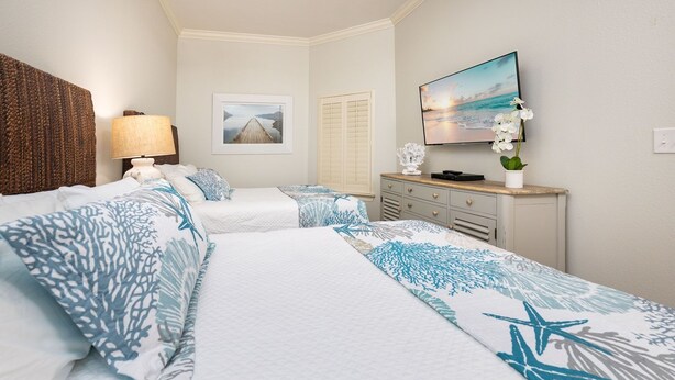 A hotel room with two beds, a nightstand, a lamp, a framed picture, a dresser, and a window with shutters. - The Oasis 904 Formerly Phoenix West Ii 904 3 Bedroom Condo (Orange Beach)