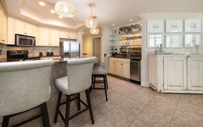 A modern kitchen with a central island, bar stools, stainless steel appliances, and decorative glass shelves. - The Oasis 904 Formerly Phoenix West Ii 904 3 Bedroom Condo (Orange Beach)