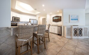 A kitchen with white cabinets, a microwave, a refrigerator, and a sink. There are wicker chairs and a dining table. - The Oasis 2509 Formerly Phoenix West Ii 2509 3 Bedroom Condo (Orange Beach)