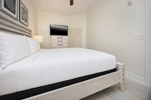 Ein ordentlich bezogenes Bett mit gesteppter weißer Decke, ein Nachttisch mit Lampe, ein flachbildfernseher über dem Bett und ein weißer Kleiderschrank mit Spiegel.