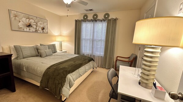A bedroom with a bed, bedside tables, a desk, and a chair. There is a window with curtains, a ceiling fan, and a wall-mounted lamp.