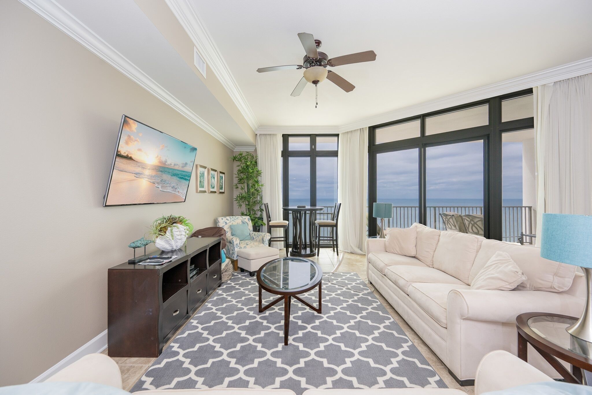 A living room with a ceiling fan, a large window with curtains, a sofa, a coffee table, and a TV mounted on the wall.