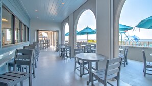A covered outdoor seating area with tables and chairs, large windows, and blue umbrellas. - The Oasis 2306 Formerly Phoenix West Ii 2306 3 Bedroom Condo (Orange Beach)