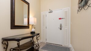 A hallway with a wooden console table, a wall-mounted mirror, a white door with a red sign, and a doormat. - The Oasis 3006 Formerly Phoenix West Ii 3006 3 Bedroom Condo (Orange Beach)