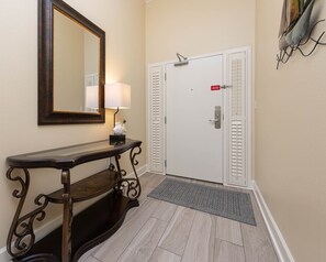A hallway with a wooden console table, a wall-mounted mirror, a white door with a red sign, and a doormat. - The Oasis 3006 Formerly Phoenix West Ii 3006 3 Bedroom Condo (Orange Beach)