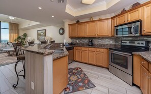 A modern kitchen with granite countertops, wooden cabinets, stainless steel appliances, and a dining area with a sofa and chairs. - The Oasis 3006 Formerly Phoenix West Ii 3006 3 Bedroom Condo (Orange Beach)