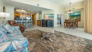 A spacious living room with a large sofa, a dining table with chairs, a kitchen area with a bar, and a painting of a beach scene. - The Oasis 3006 Formerly Phoenix West Ii 3006 3 Bedroom Condo (Orange Beach)