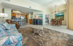 A spacious living room with a large sofa, a dining table with chairs, a kitchen area with a bar, and a painting of a beach scene. - The Oasis 3006 Formerly Phoenix West Ii 3006 3 Bedroom Condo (Orange Beach)