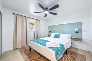 A hotel room with a bed, a ceiling fan, a window with curtains, and a nightstand with a lamp. - Big Island Kahaluu Surf Boutique #4 2 Bedroom Apts (Kailua-Kona)